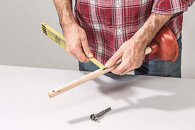 Stiel eines Pömpels wird mit einem Zollstock ausgemessen, um daraus einen Haken für den Raumteiler aus Birkenstämmen zu bauen