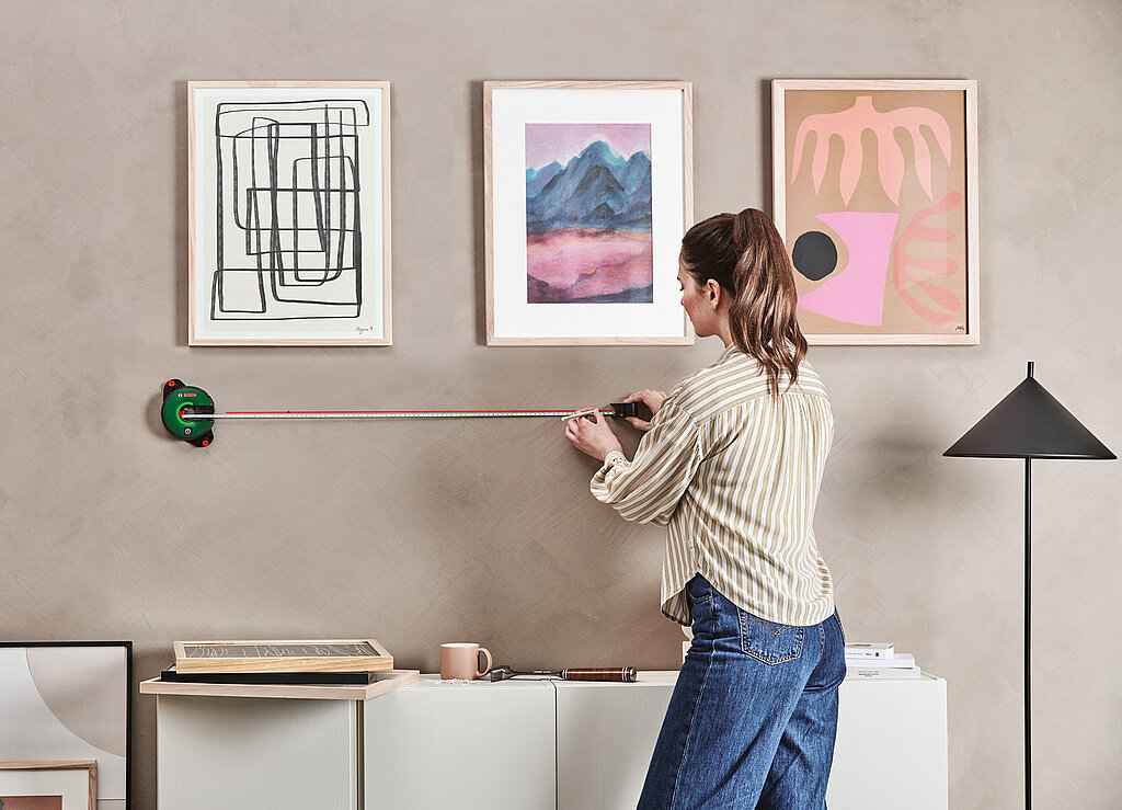 3 Bilder hängen über einem Sideboard, Frau misst Abstände mit einem Linienlaser