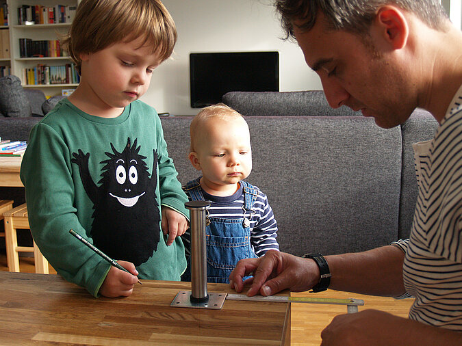 Tipps zum Heimwerken mit Kindern: Zwei kleine Kinder helfen beim Anzeichnen und Vorbereiten eines DIY-Projekts, während ein Erwachsener eine Tischstütze positioniert – spielerisches Lernen durch gemeinsames Handwerken.