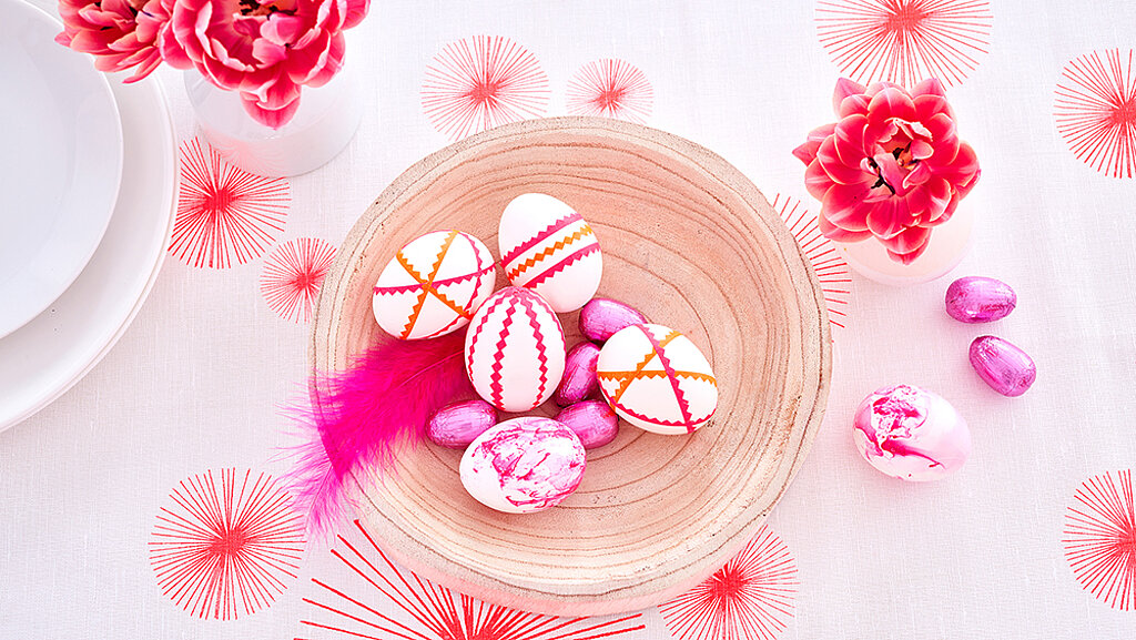 Holzschale mit Neon-Ostereiern, pinker Feder und pinken Schokoeiern auf weißer Tischdecke mit roten Sternmustern, daneben rosa marmoriertes Ei und pinke Blumen in Vasen