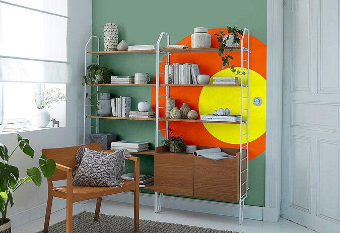 Raumgestaltung Wohnzimmer kreativ – Regalwand mit farbigen Akzenten in Orange und Gelb, kombiniert mit Sessel, Pflanzen und dekorativen Wohnaccessoires