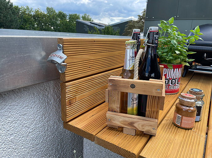 Grillplatz für die Terrasse bauen: Integrierter Flaschenöffner an der Holzarbeitsfläche, daneben ein DIY-Flaschenträger mit Bier, frischen Kräutern und Grillgewürzen – ideal für entspannte Abende auf der Terrasse