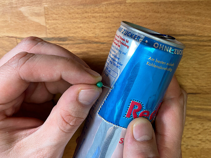 Detailbild, mit einer Stecknadel wird ein Loch am unteren Rand einer Red-Bull-Dose gestochen – Vorbereitung für die Lichterkettenöffnung.“
