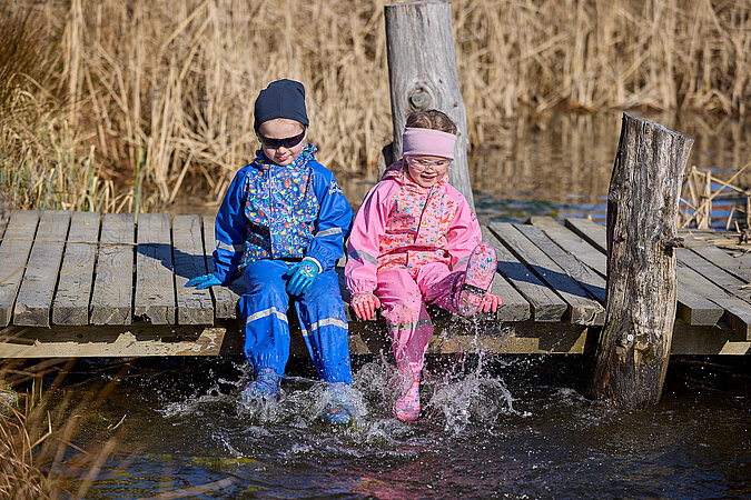 Tipps zum Heimwerken mit Kindern: Zwei Kinder in wetterfester Kleidung sitzen auf einem Holzsteg und plantschen im Wasser – spielerische Erkundung der Natur als Teil von Outdoor-DIY-Aktivitäten.