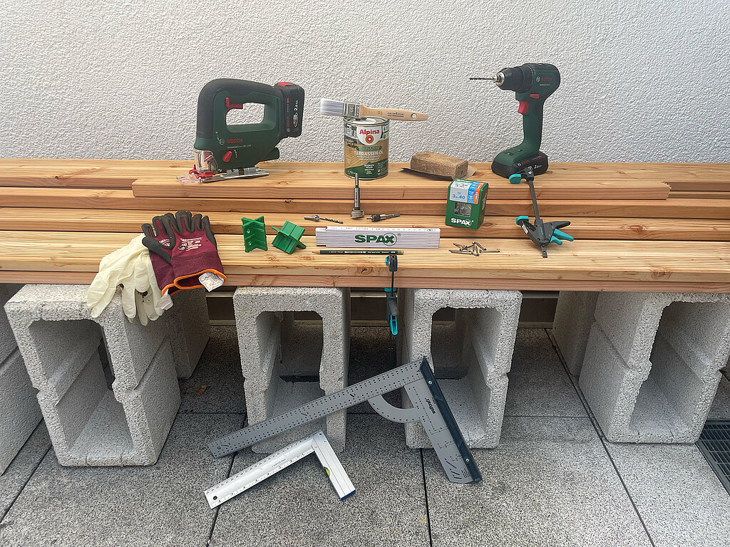 Grillplatz für die Terrasse bauen: Alle benötigten Materialien und Werkzeuge wie Stichsäge, Akkuschrauber, Holzlatten, Schrauben, Schutzhandschuhe und Betonsteine übersichtlich aufgestellt für den Start eines DIY-Projekts im Außenbereich.