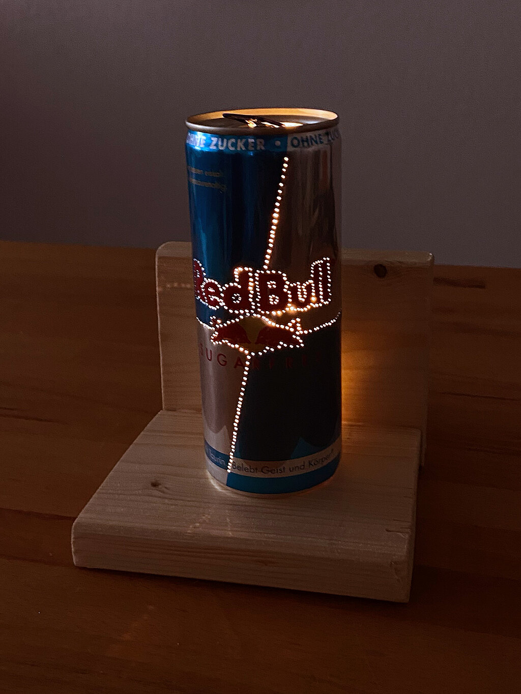 Leuchtende DIY-Tischampe aus gelochter Red-Bull-Getränkedose auf einem L-förmigen Holzpodest in dunkler Umgebung.