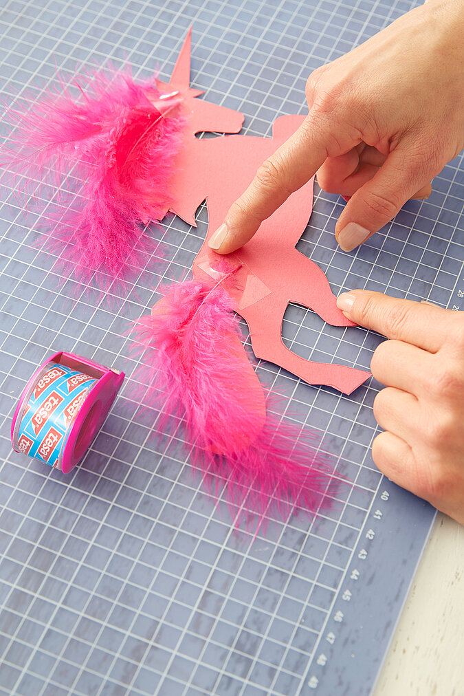 An einem rosa, glitzernden Einhorn wird ein Schweif aus pinken Federn angeklebt