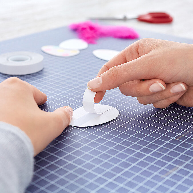 Hände kleben einen Papierstreifen als Abstandshalter mittig auf ein ausgeschnittenes Ei, im Hintergrund liegen weitere Ei-Formen und pinke Federn