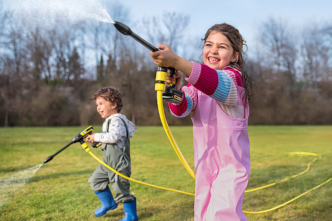 Tipps zum Heimwerken mit Kindern: Mädchenund Junge stehen auf einer Wiese und spritzen mit einem Kinder-Reinigungsgerät 