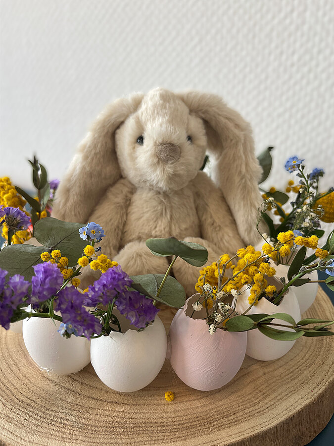 Osterkranz aus Eierschalen basteln als fertige Osterdeko mit Blumen, bunten Eierschalen und einem kleinen Stoffhasen auf einer Holzscheibe