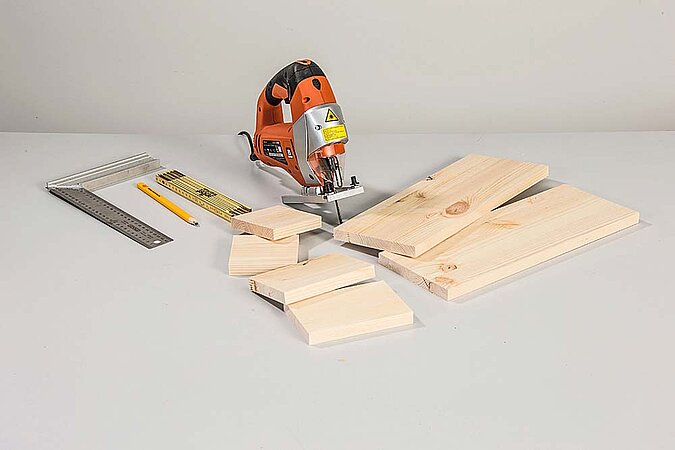 Stichsäge, Winkel, Zollstock, Bleistift und zugeschnittene Holzteile für kleine Regale am Raumteiler aus Birkenstämmen liegen auf einer Werkbank