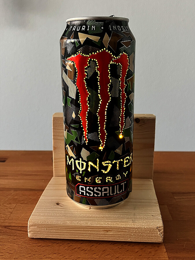 Leuchtende DIY-Tischampe aus gelochter Monster-Energy-Getränkedose auf einem L-förmigen Holzpodest in etwas hellerer Umgebung.