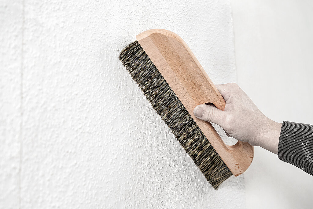 Wände richtig tapezieren – praktische Tipps zur Vorbereitung: Hand mit Tapezierbürste glättet sorgfältig die strukturierte Wandoberfläche als Vorbereitung für das Tapezieren.