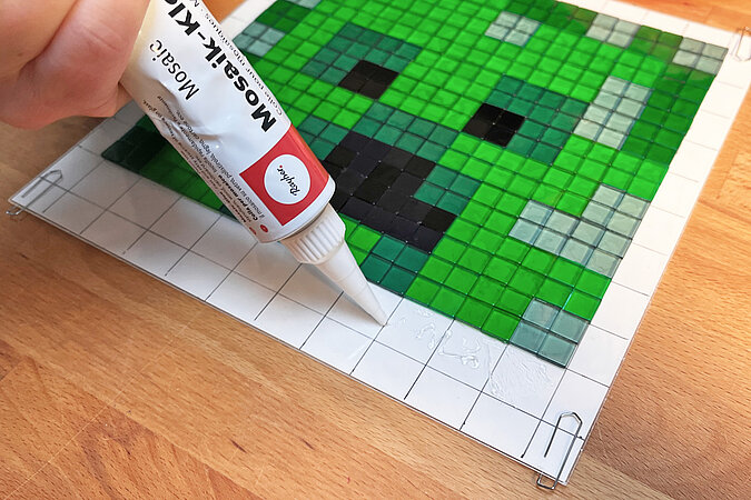 Creeper-Deko-Leuchte basteln beim gezielten Verkleben dunkler Mosaiksteine für Augen und Mund des Creepers