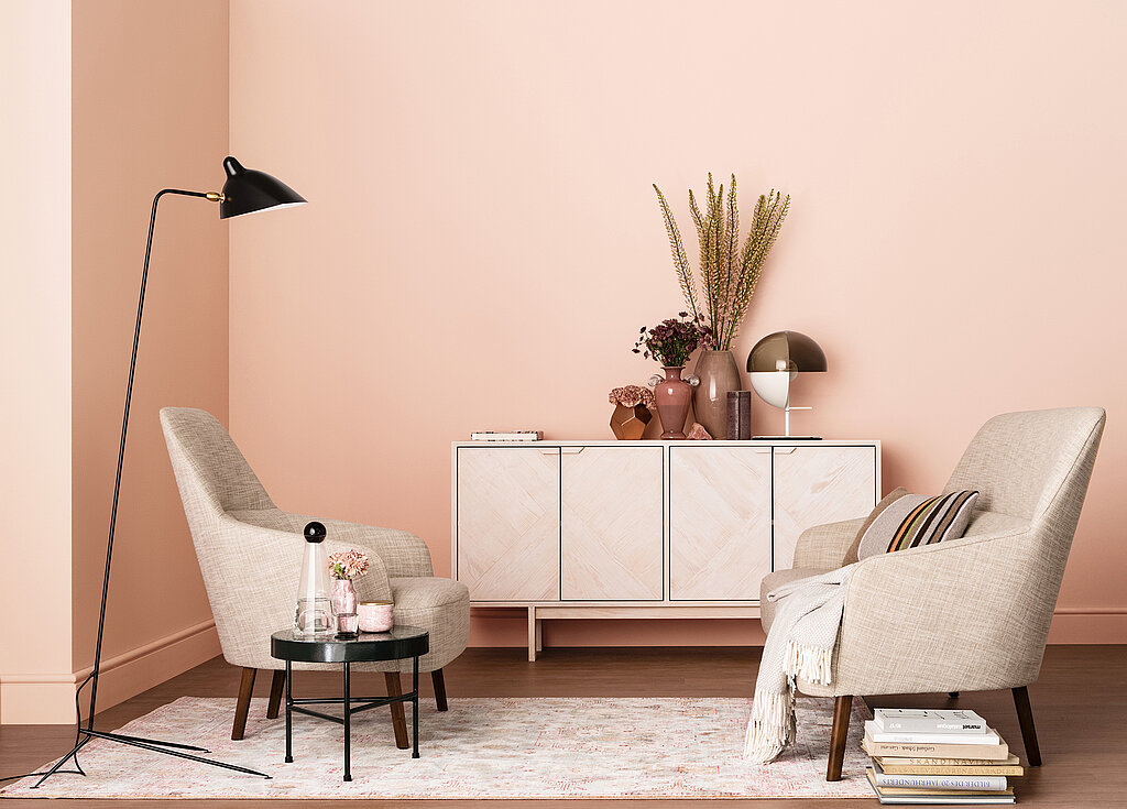 Raumgestaltung Wohnzimmer elegant – helle Möbel mit Sideboard, Sessel und Stehlampe vor zart rosafarbener Wand im modernen Wohnstil