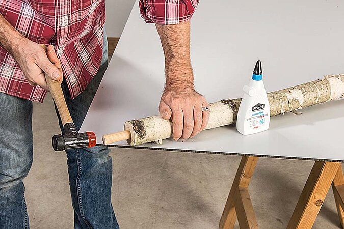Holzdübel wird in einen Birkenstamm für einen Raumteiler aus Birkenstämmen eingeleimt und mit einem Hammer eingeschlagen