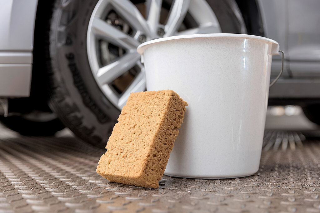 Auto selbst waschen Zubehör – Eimer mit Wasser und Schwamm vor Autoreifen, grundlegende Utensilien für die gründliche Handwäsche und Lackpflege im eigenen Hof.