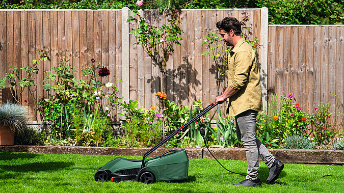 Akku Gartengeräte im Überblick – Mann mäht mit kabellosem Akku-Rasenmäher den gepflegten Gartenrasen vor Holzzaun und blühenden Beeten für flexible und leise Gartenarbeit