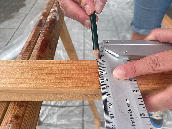 Grillplatz für die Terrasse bauen: Präzises Anzeichnen mit Metallwinkel und Bleistift auf einer Holzlatte zur Vorbereitung des Zuschnitts – wichtige Vorarbeit für den stabilen Aufbau einer DIY-Grillstation.