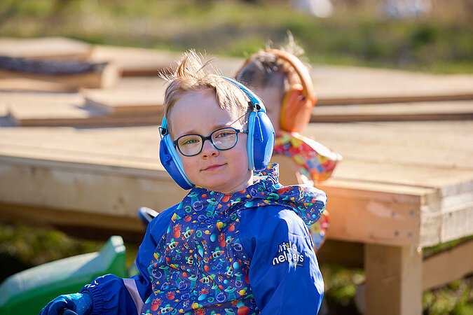 Tipps zum Heimwerken mit Kindern: Junge mit Gehörschutz und bunter Arbeitskleidung sitzt auf einem Trettraktor vor einer Holzbaustelle – spielerisches Heranführen an sicheres Arbeiten im DIY-Bereich.