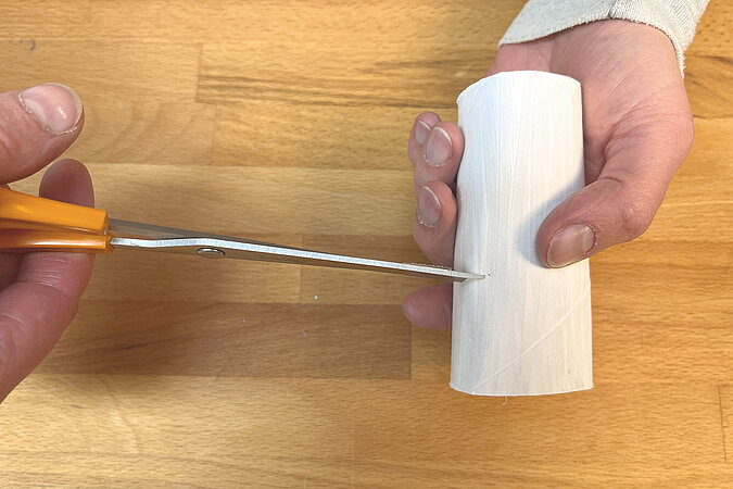 eine Hand schneidet mit einer Schere eine Oeffnung fuer einen Schnabel in einen leeren Toilettenrollenkern