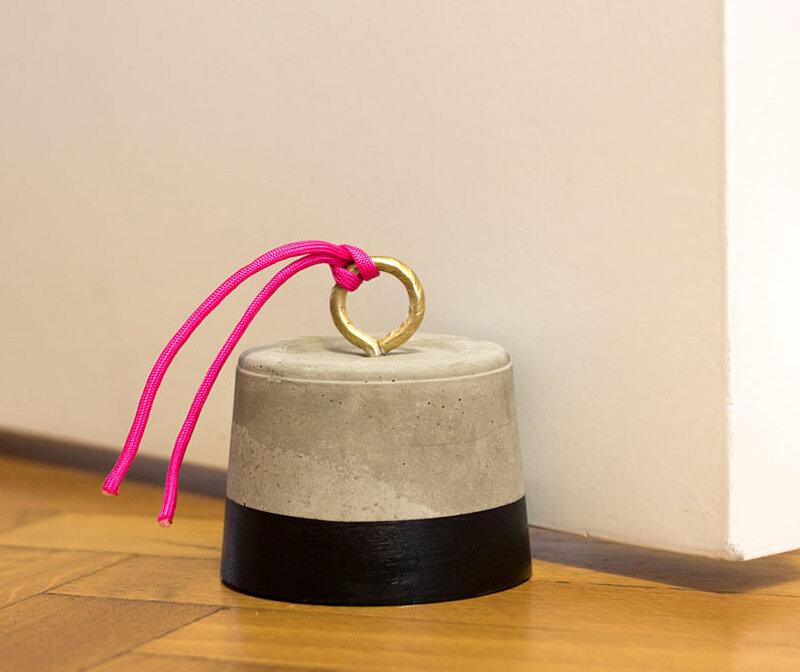 Türstopper aus Beton selber machen – schlichter DIY-Türstopper aus gegossenem Beton mit schwarzem Sockel, pinkfarbener Schnur und Messingring, der eine weiße Tür auf Holzfußboden fixiert