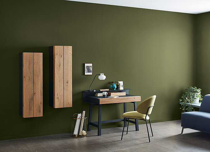 Raumgestaltung Arbeitszimmer modern – Schreibtisch mit Stuhl und Dekoration vor dunkelgrüner Wand mit minimalistischen Holzelementen und stilvollem Design
