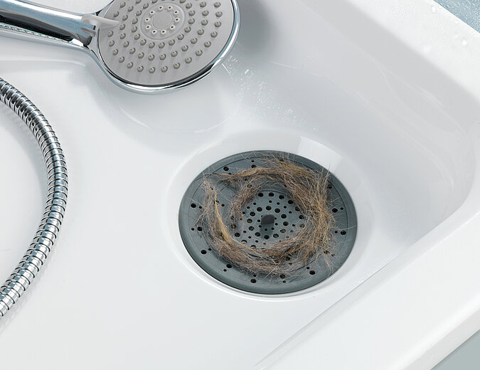 Ein Duschkopf liegt in eine Dusche daneben ein Haarsieb mit Haaren auf dem Abfluss