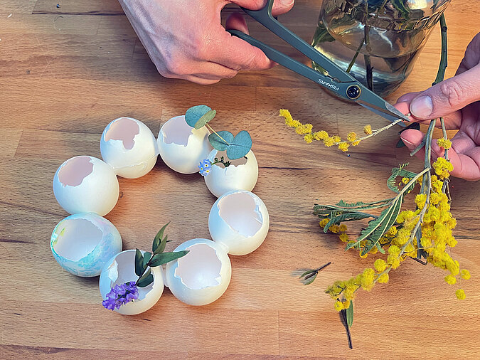 Osterkranz aus Eierschalen basteln mit Heißkleber und frischen Blumen, während die Eierschalen zu einem dekorativen Kranz arrangiert werden