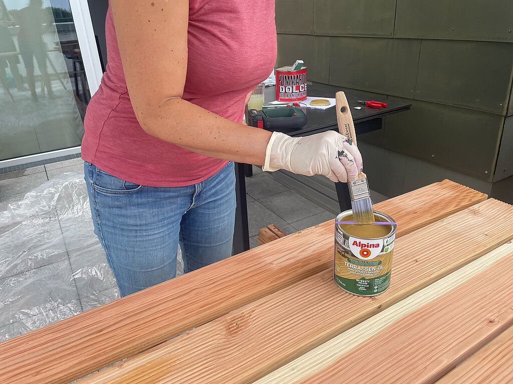 Grillplatz für die Terrasse bauen: Terrassenöl wird mit dem Pinsel aus der Dose entnommen – Vorbereitung zur Holzpflege und Versiegelung der Dielen für langlebige und wetterfeste DIY-Grillmöbel.
