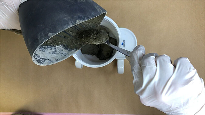 Türstopper aus Beton selber machen – flüssiger Beton wird aus einem Mischbecher mithilfe eines Löffels in eine Silikonform gegossen, präziser DIY-Arbeitsschritt auf Werkunterlage