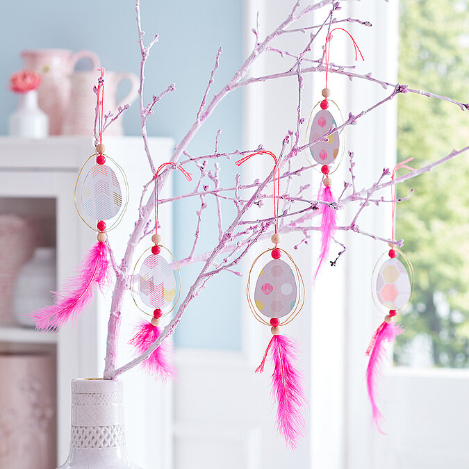 Hellrosa bemalte Zweige in weißer Vase mit fünf selbstgemachten Ei-Anhängern aus gemustertem Papier im goldenen Drahtrahmen, mit Holzperlen und pinken Federn vor einem großen Fenster
