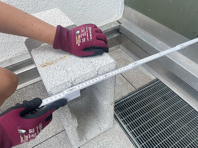 Grillplatz für die Terrasse bauen: Ausmessen und Positionieren eines Betonsteins mit Zollstock – exakte Vorbereitung für den stabilen Unterbau der selbstgebauten Grillstation im Außenbereich.