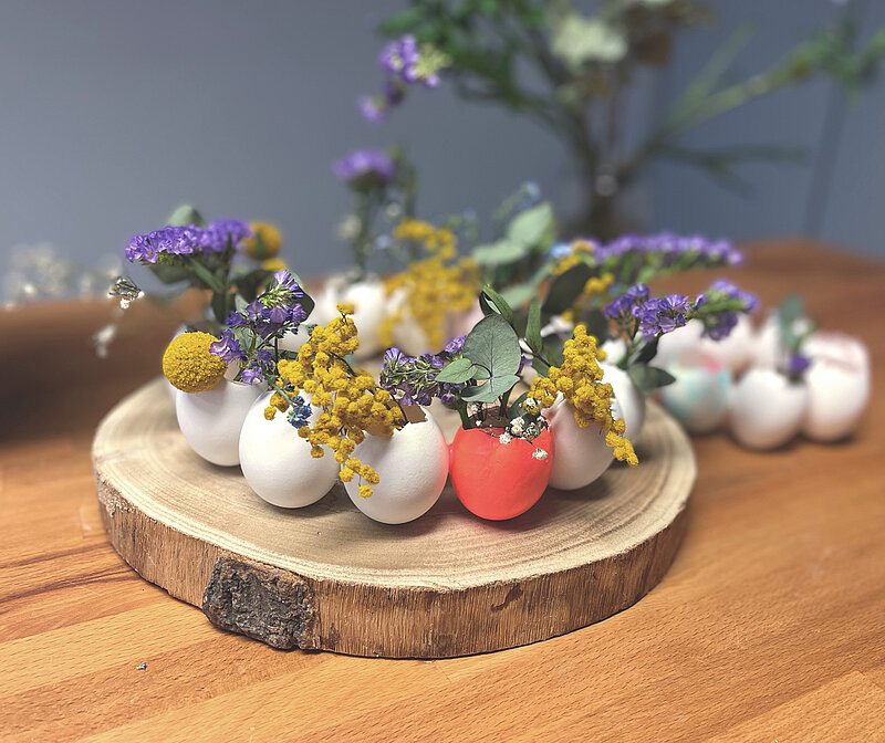 Osterkranz aus Eierschalen basteln mit bemalten Eierschalen, Trockenblumen und frischen Blüten auf einer runden Holzscheibe als frühlingshafte Tischdeko zu Ostern