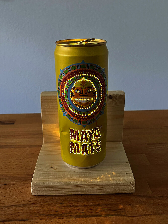 Leuchtende DIY-Tischampe aus gelochter Maya-Mate-Getränkedose auf einem L-förmigen Holzpodest in dunkler Umgebung.