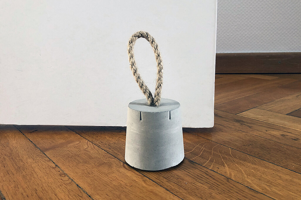 Türstopper aus Beton selber machen – schlichter, selbstgemachter Beton-Türstopper mit Kordelgriff hält eine weiße Tür auf Holzfußboden offen, minimalistisches DIY-Wohnaccessoire