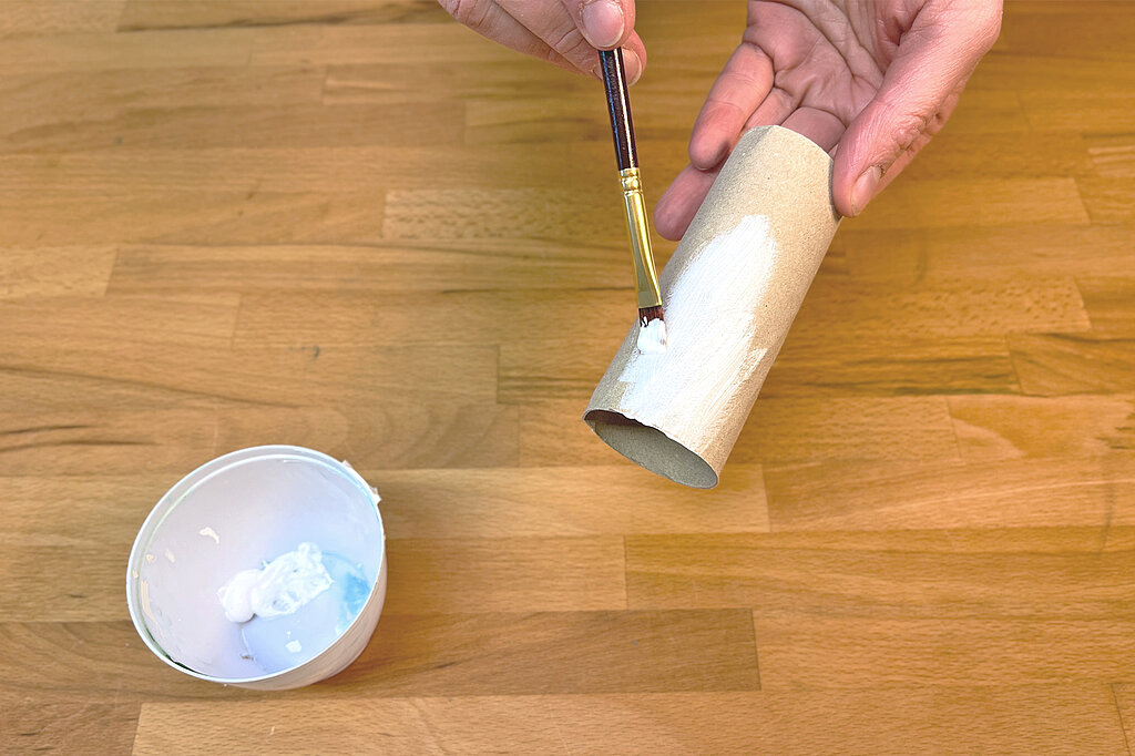 ein leerer Pappkern einer Toilettenrolle wird mit einem Pinsel weiss angemalt