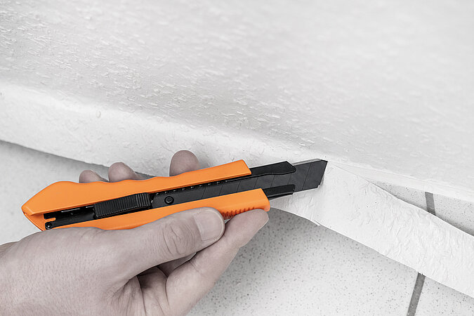 Wände richtig tapezieren – praktische Tipps für saubere Abschlüsse: Hand schneidet überstehende Tapete mit Cuttermesser exakt an der Fußleiste entlang für einen präzisen Wandabschluss.