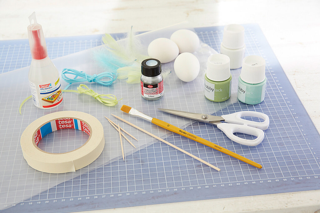 Osterstrauch DIY Materialien wie Acrylfarben, ausgeblasene Eier, Pinsel, Klebeband und Schere auf Bastelunterlage für kreative Osterdeko