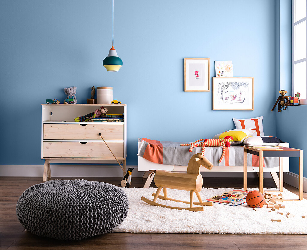 Raumgestaltung Kinderzimmer kreativ – helles Zimmer mit blauer Wand, Kinderbett, Kommode, Schaukelpferd und verspielten Dekorationselementen