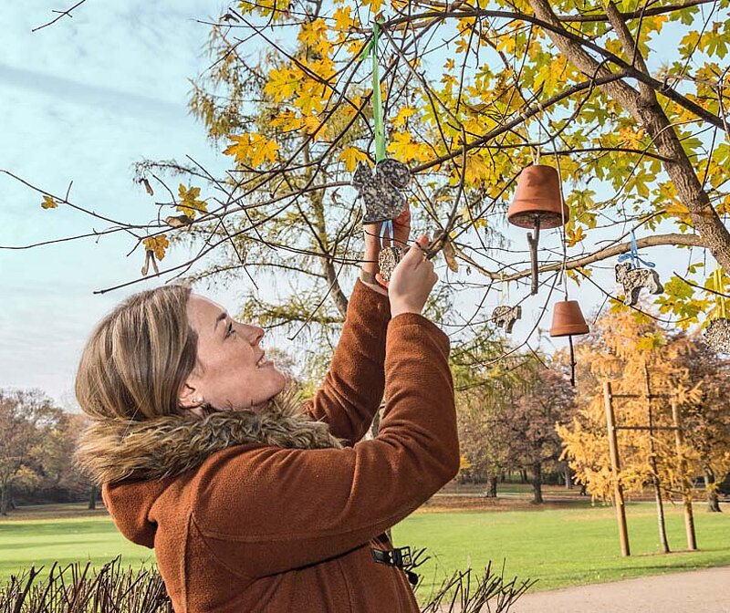 Vogelfutter selber machen im Herbst – Frau hängt selbstgemachte Futteranhänger und Tontöpfe mit Vogelfutter an einen Baum im Garten