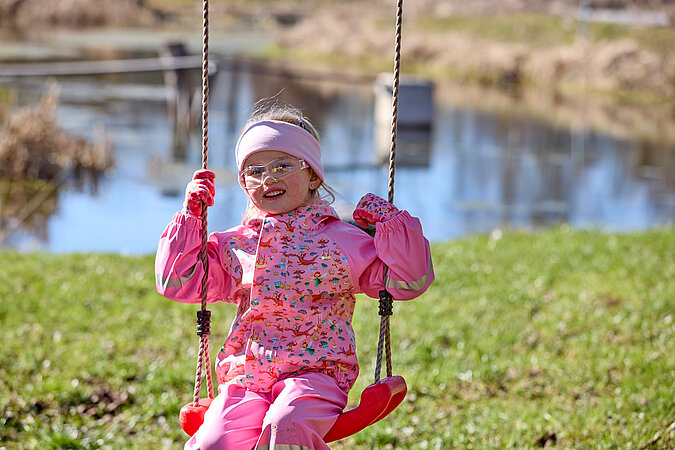 Tipps zum Heimwerken mit Kindern: Mädchen in bunter Outdoor-Kleidung schaukelt fröhlich vor einem Teich – selbstgebaute Schaukel als kreatives DIY-Projekt im Garten.
