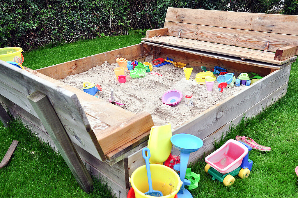 Sandkasten mit seitlichen Klappdeckeln und Spielsachen wie Eimer und Schäufelchen