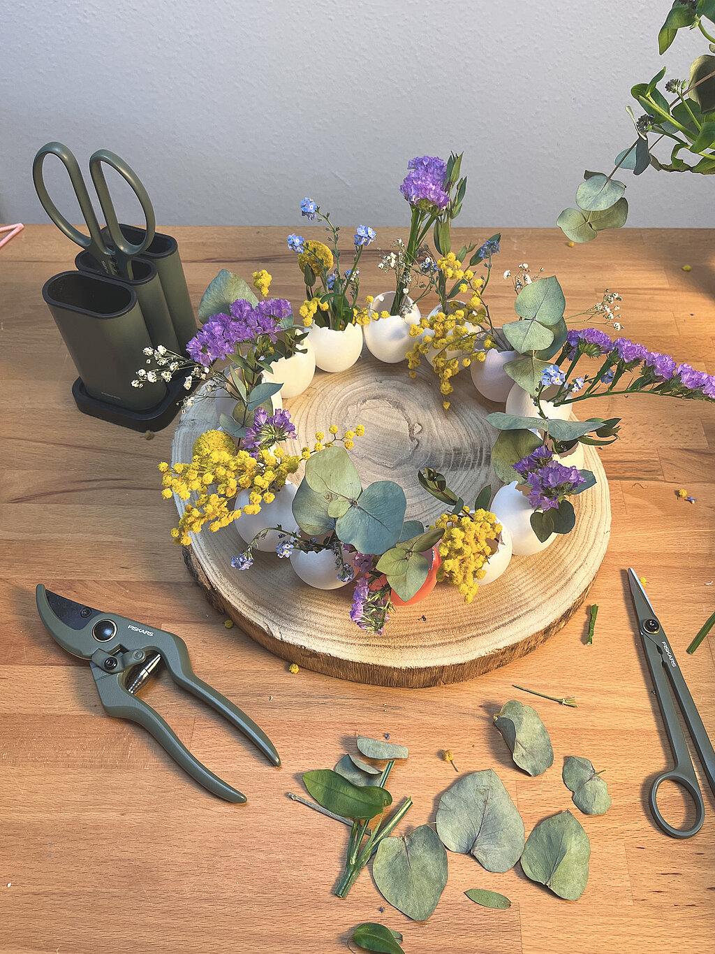 Osterkranz aus Eierschalen basteln Schritt-für-Schritt mit Eierschalen, Eukalyptus, Trockenblumen und Gartenschere auf einer Holzscheibe als kreative DIY-Osterdeko