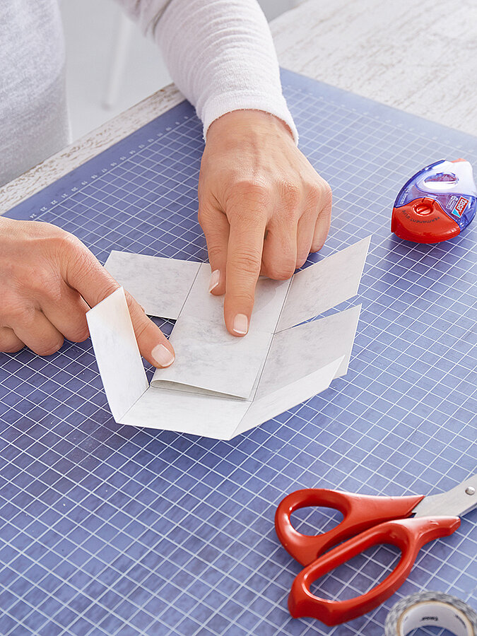 Hände flechten und drücken Papierstreifen zu einer kleinen Schachtel, rote Schere und tesa Kleberoller liegen auf der Schneidematte