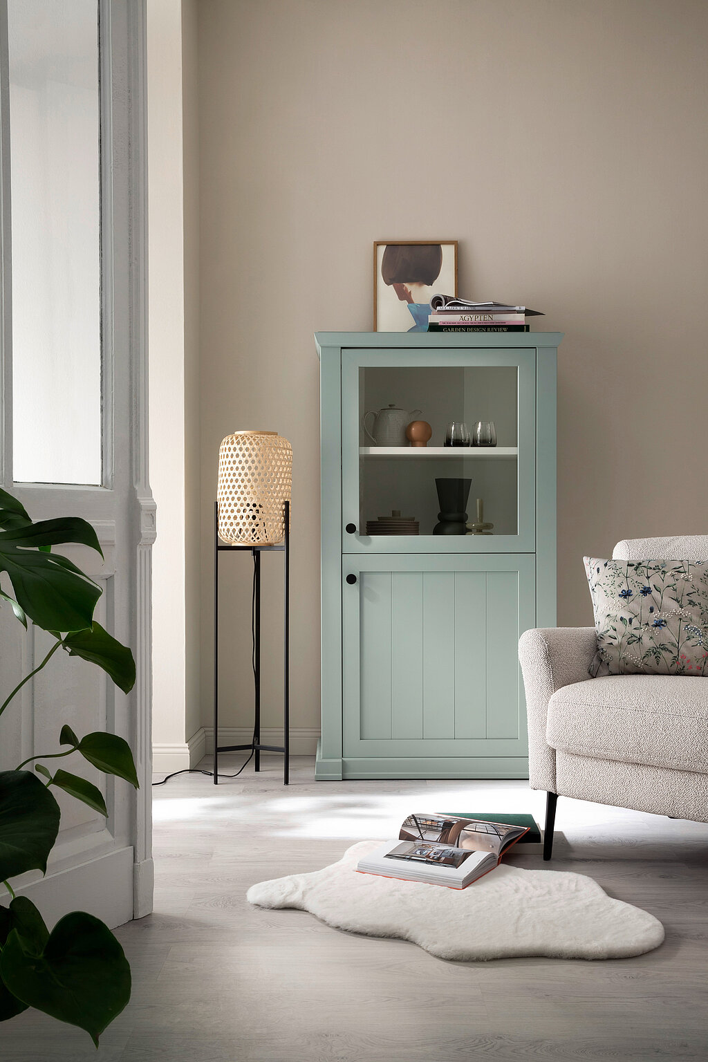 Raumgestaltung Wohnzimmer stilvoll – mintfarbene Vitrine mit Dekoration, Stehlampe und Sessel vor neutraler Wand für ein modernes Wohnkonzept