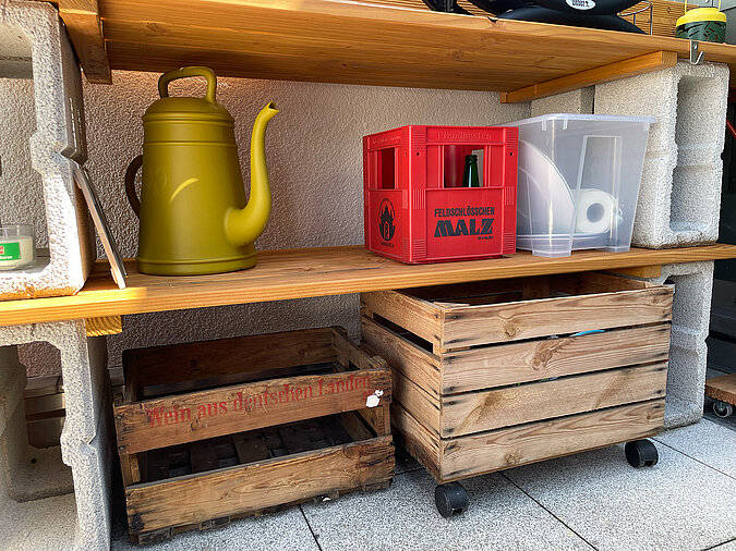 Grillplatz für die Terrasse bauen: Untere Ablageflächen der DIY-Grillstation mit Holzkisten auf Rollen, einer roten Getränkekiste, transparenter Aufbewahrungsbox und stylischer Gießkanne – clevere Stauraumlösungen für die Terrasse