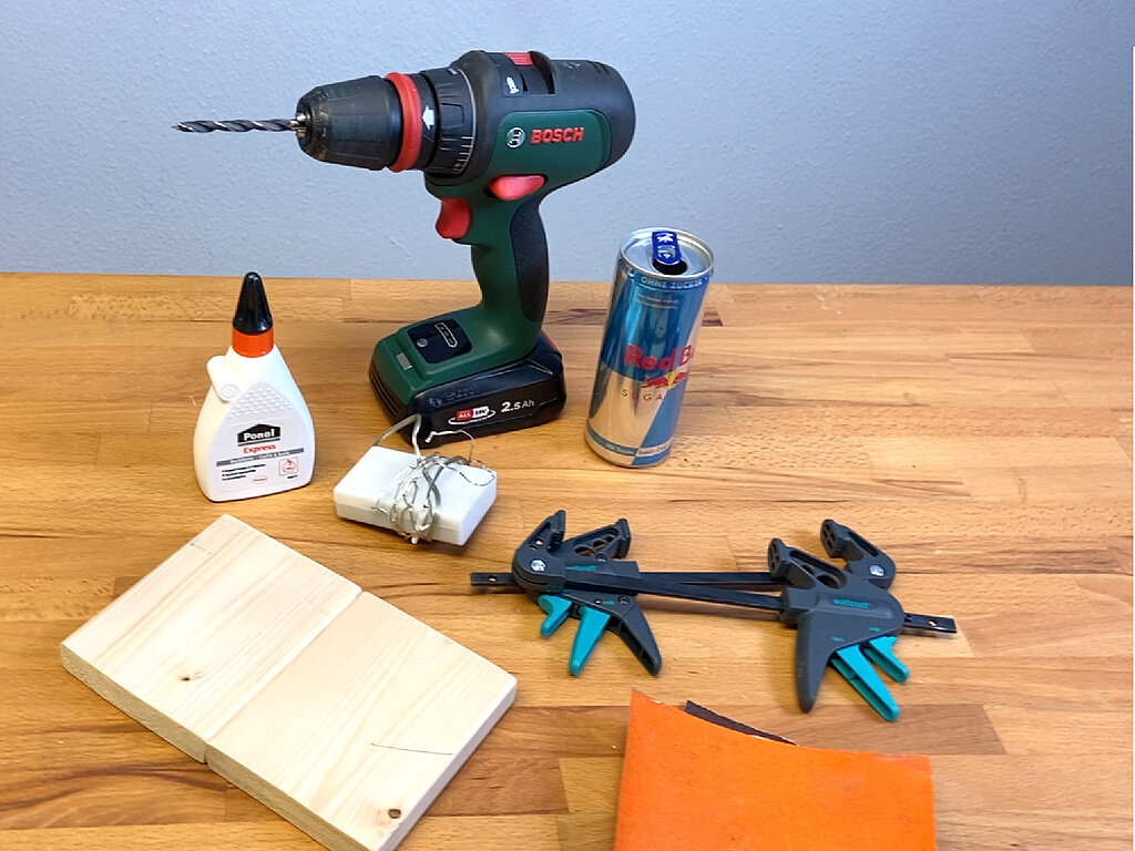 Materialien und Werkzeuge für eine DIY LED-Tischleute aus Getränkedose: Bohrmaschine, Red-Bull-Dose, Holzstücke, Lichterkette mit Batteriefach, Holzleim, Schraubzwingen und Schleifpapier.