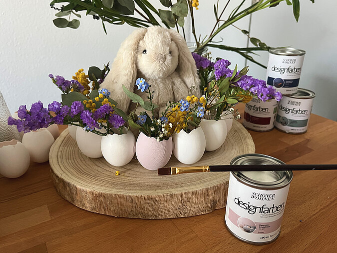 Osterkranz aus Eierschalen basteln als stilvolle Osterdeko mit bemalten Eierschalen, bunten Frühlingsblumen und Deko-Hasen auf einer runden Holzscheibe