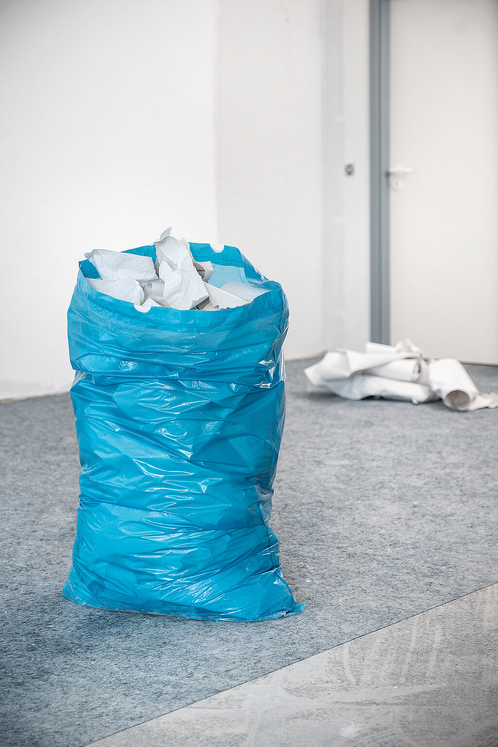 Wände richtig tapezieren – praktische Tipps zur Entsorgung: Blauer Müllsack gefüllt mit alten Tapetenresten steht auf dem Boden, im Hintergrund weitere entfernte Tapetenbahnen – Aufräumen nach dem Tapezieren.